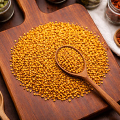 A heap of small, round golden-yellow bee pollen granules resting on a dark wooden board next to a wooden spoon.